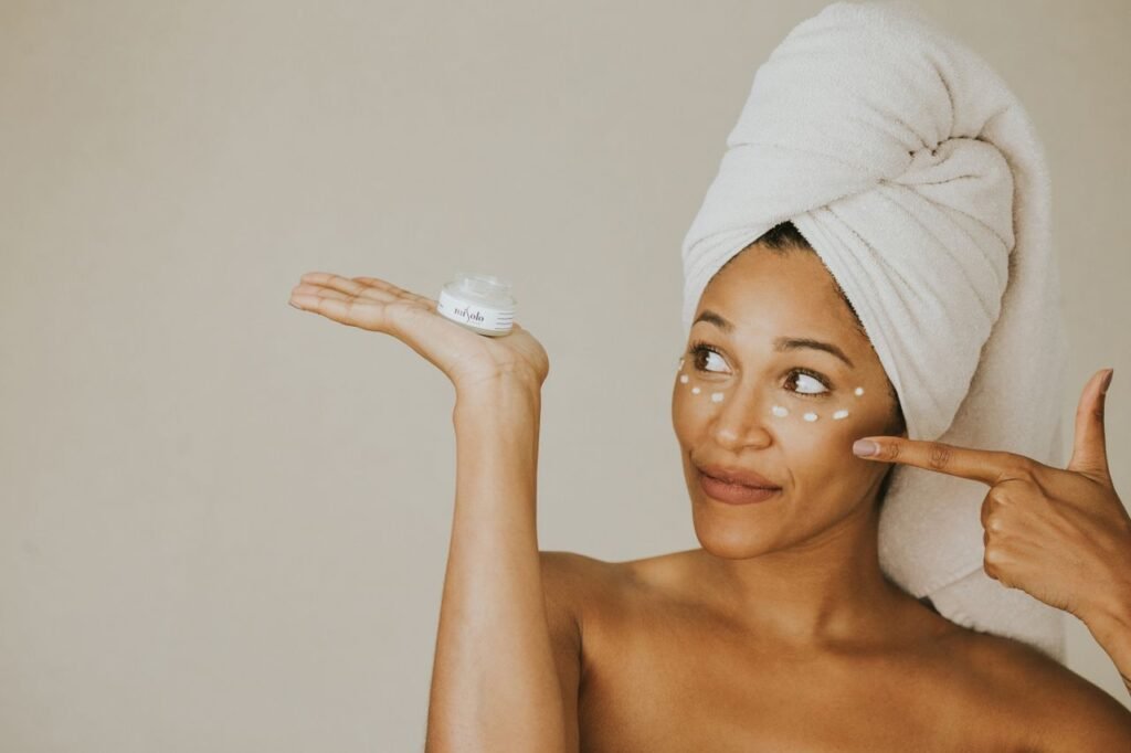 A woman applying face cream with a wrapped towel on her head, smiling.